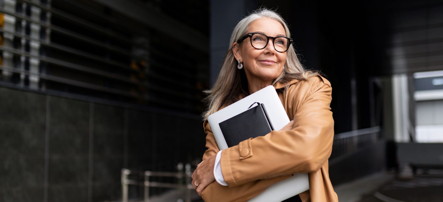 Ältere Fachfrau mit Brille hält Laptop und Unterlagen vor einem Bürogebäude