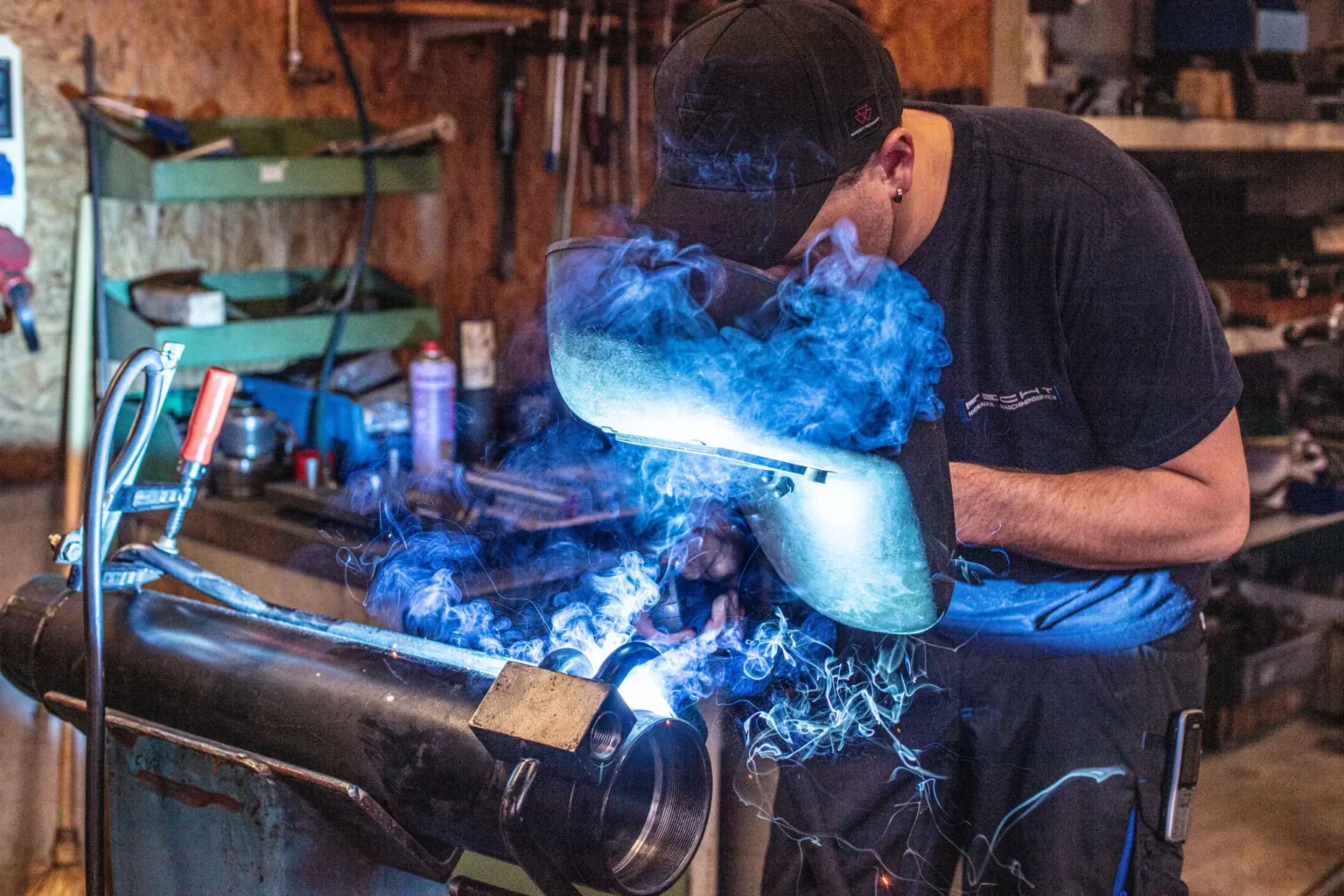 Mitarbeiter mit Schweißschirm schweißt an einem Hydraulikzylinder, blauer Rauch steigt auf